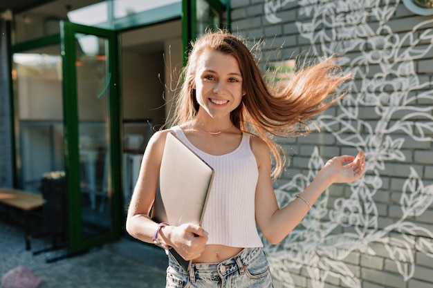 Foto grátis garota sorridente atraente feliz com laptop andando na cidade em um dia bom de sol. aluno vai para o estudo