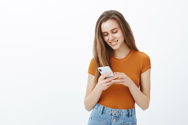 Garota sorridente atraente usando telefone celular, mensagem de texto, download de aplicativo ou assistindo vídeo
