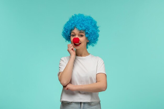 Garota sorridente do dia da mentira pensando em tocar o cotovelo com nariz vermelho em uma fantasia de palhaço cabelo azul