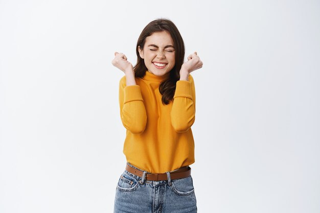 Garota sorridente e animada vencendo e comemorando, aperte as mãos alegres e pulando de alegria, triunfando, alcance objetivo ou prêmio, parede branca