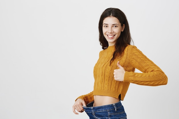 Garota sorridente e satisfeita perdeu peso, mostrando jeans soltos e polegar para cima