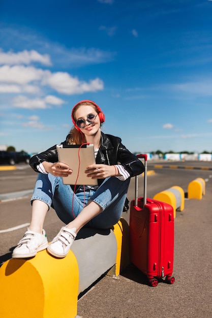 Garota sorridente em óculos de sol e fones de ouvido ouvindo música alegremente no tablet com mala vermelha perto da estrada do aeroporto