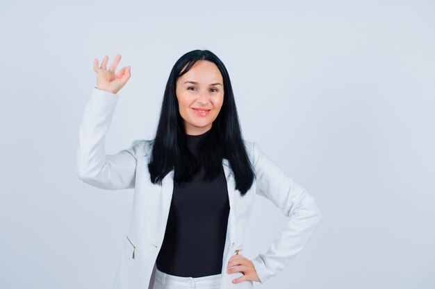 Garota sorridente está mostrando um gesto bem e colocando a outra mão na cintura em fundo branco