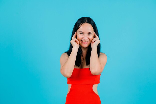 Garota sorridente está olhando para a câmera segurando o dedo indicador nas orelhas em fundo azul