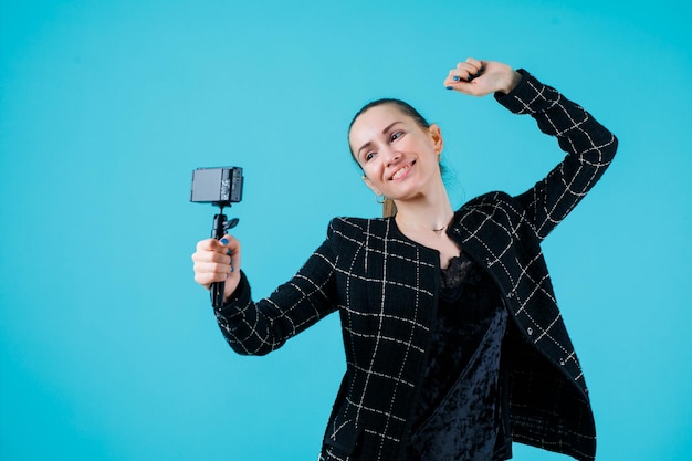 Garota sorridente está tirando selfie com sua câmera levantando o punho no fundo azul