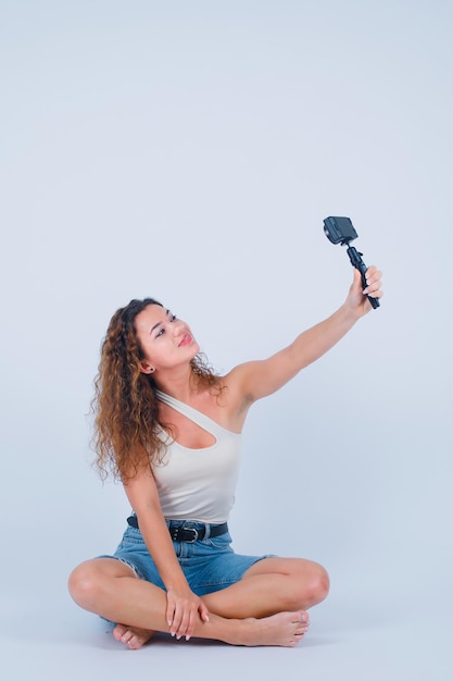 Garota sorridente está tirando selfie com sua mini câmera sentada no chão no fundo branco