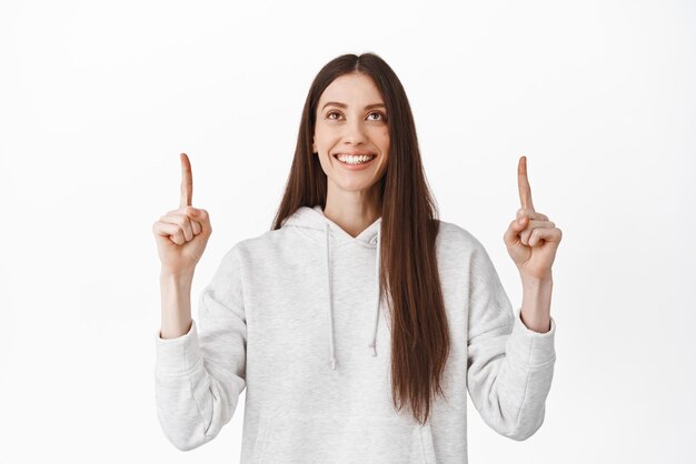 Garota sorridente sonhadora assistindo algo bonito acima apontando e olhando para cima com cara feliz promovendo produto mostrando fundo branco de anúncio