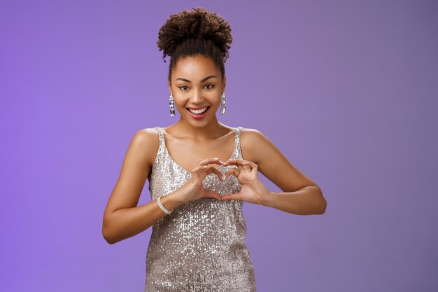 Garota totalmente amada adora olhar deisgners gratos espreitar vestido elegante festa de aniversário mostrar gesto de coração sorrindo alegremente expressando amor, sentimentos apaixonados comoventes em pé de bom grado fundo azul.