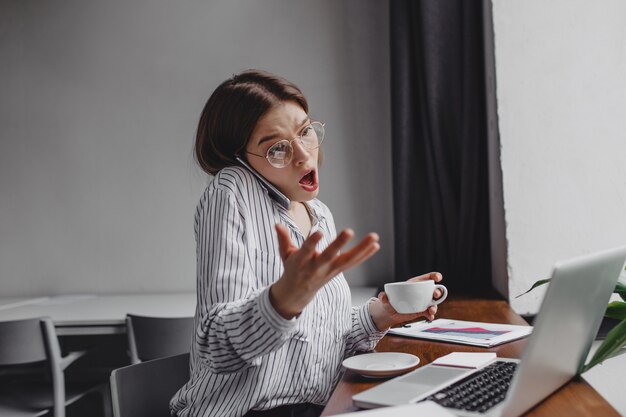 Garota trabalhadora em copos, emocionalmente e indignadamente falando no telefone, sentado no escritório com o laptop e uma xícara de café.