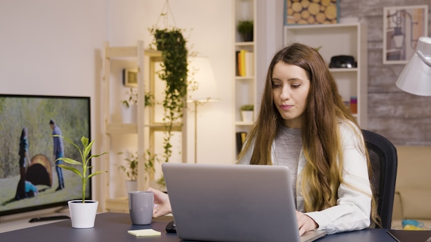 Garota trabalhando no laptop do escritório em casa. Menina tomando um gole de café.