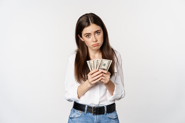 Foto grátis garota triste triste contando dólares de dinheiro e de mau humor, de pé em roupas casuais contra fundo branco