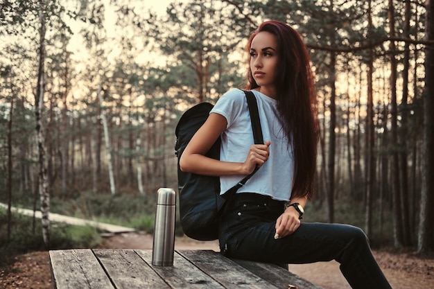 Garota turista sensual sentada em uma mesa de madeira, fazendo uma pausa em uma bela floresta de outono ao pôr do sol.