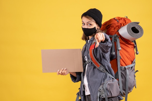 garota viajante com máscara preta e mochila segurando um papelão apontando para a câmera