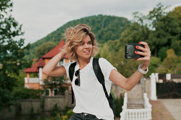 Garota viajante sorridente feliz com penteado encaracolado, fazendo selfie sobre a montanha. Viajante usando celular de mão feminina. Turismo na montanha, estilo de vida de verão