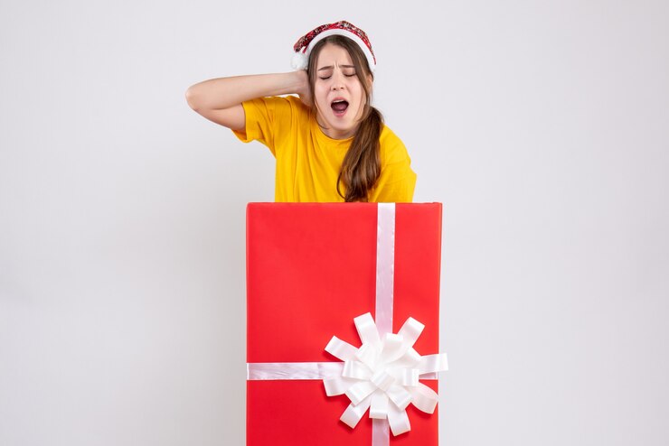 Foto grátis garota zangada de frente com chapéu de papai noel segurando a cabeça em pé atrás de um grande presente de natal