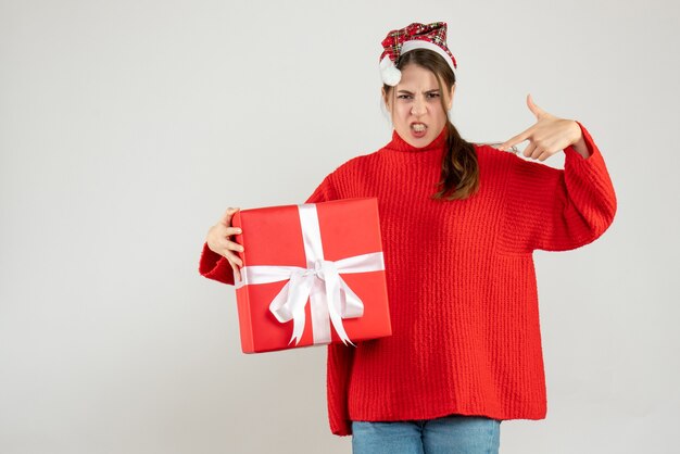 Garota zangada de frente com chapéu de Papai Noel segurando o dedo do presente e apontando para si mesma