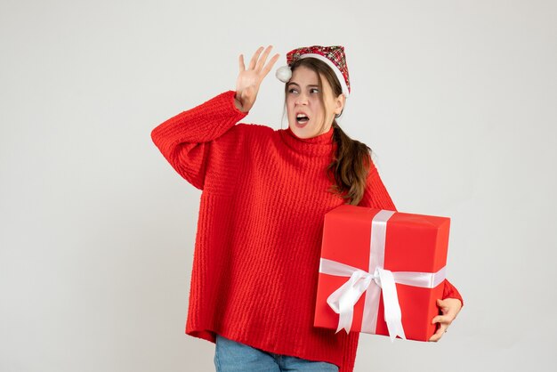 Garota zangada de frente com chapéu de Papai Noel segurando um presente de pé