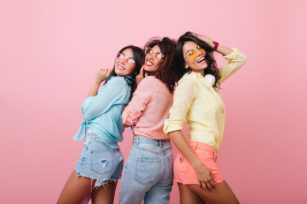 Garotas despreocupadas em camisas de algodão coloridas posando juntas e sorrindo. Retrato interior de mulheres atraentes que expressam emoções felizes.