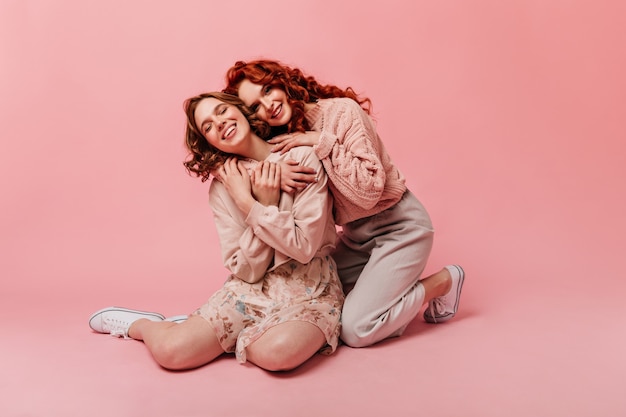 Garotas entusiasmadas sentadas no chão com um sorriso. foto de estúdio de mulheres bonitas rindo sobre fundo rosa.