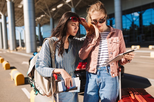 Garotas legais em óculos de sol segurando passaporte com bilhetes enquanto pensativamente usando o tablet junto com o aeroporto em segundo plano