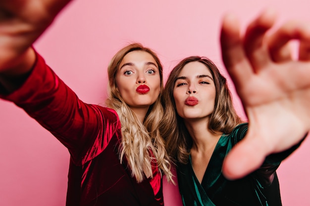 Garotas lindas em vestidos de veludo posando com expressão facial de beijo. Tiro interno de senhoras elegantes despreocupadas fazendo selfie.