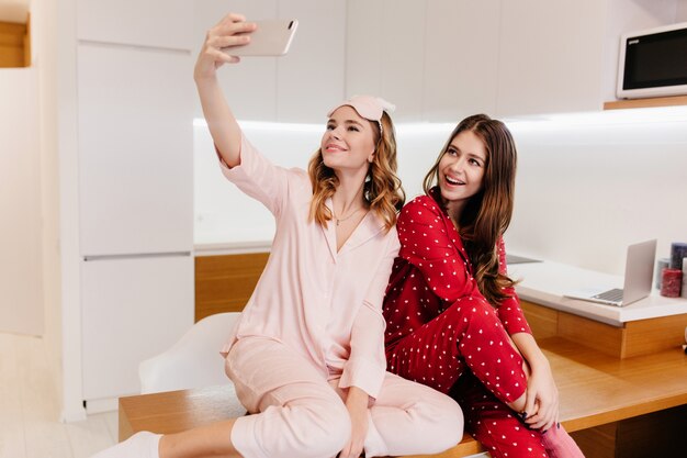 Garotas românticas sentadas juntas na mesa de madeira e tirando fotos de si mesmas. Foto interna de lindas senhoras de pijama fazendo selfie na cozinha.