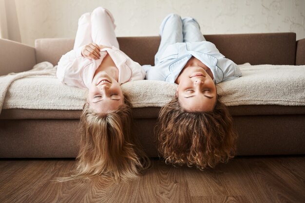 Garotas só querem se divertir. Namoradas bonitas, deitado no sofá de cabeça para baixo com o cabelo tocando o chão, vestindo roupas de dormir aconchegantes, sorrindo e relaxando com os olhos fechados, passando muito tempo juntos.