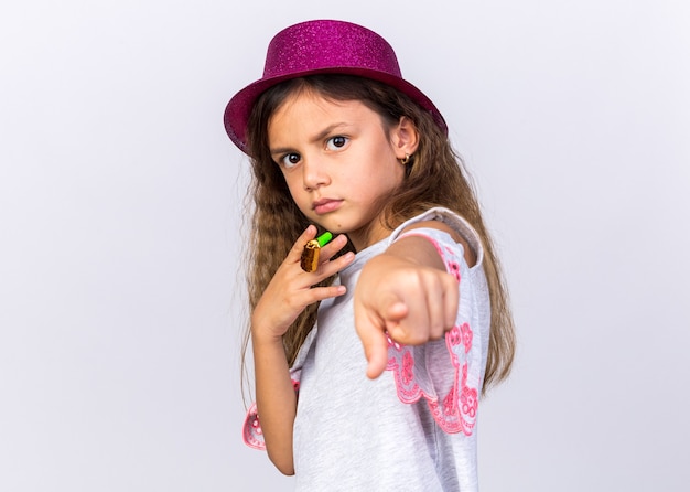 Garotinha caucasiana confiante com chapéu de festa roxo segurando o apito de festa e apontando isolado na parede branca com espaço de cópia