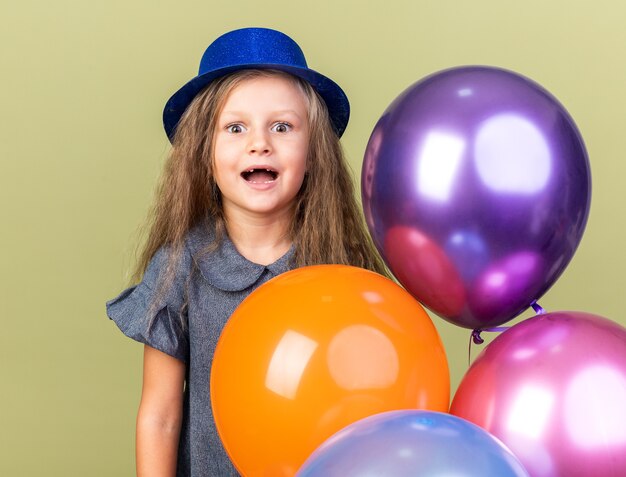 garotinha loira animada com chapéu de festa azul segurando balões de hélio isolados na parede verde oliva com espaço de cópia