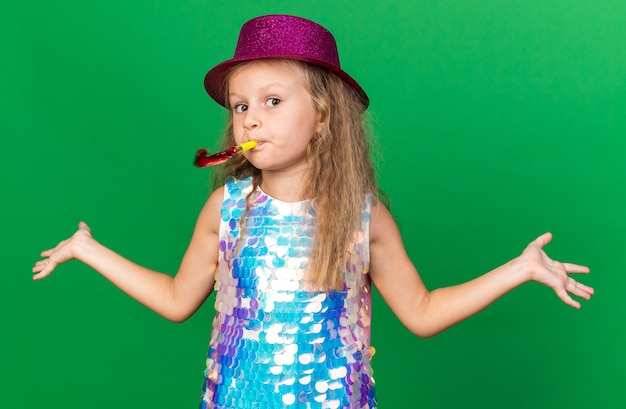 garotinha loira confusa com chapéu de festa roxo, soprando apito de festa e mantendo as mãos abertas, isolado na parede verde com espaço de cópia