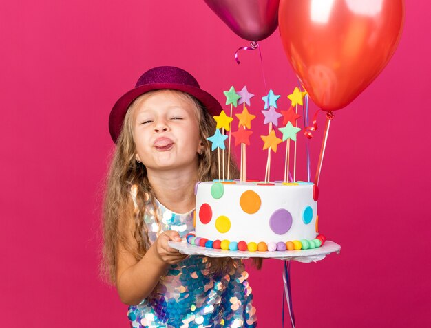 garotinha loira engraçada com chapéu de festa roxo enfia a língua para fora segurando balões de hélio e um bolo de aniversário isolado na parede rosa com espaço de cópia