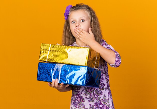 garotinha loira surpresa colocando a mão na boca e segurando caixas de presente isoladas na parede laranja com espaço de cópia