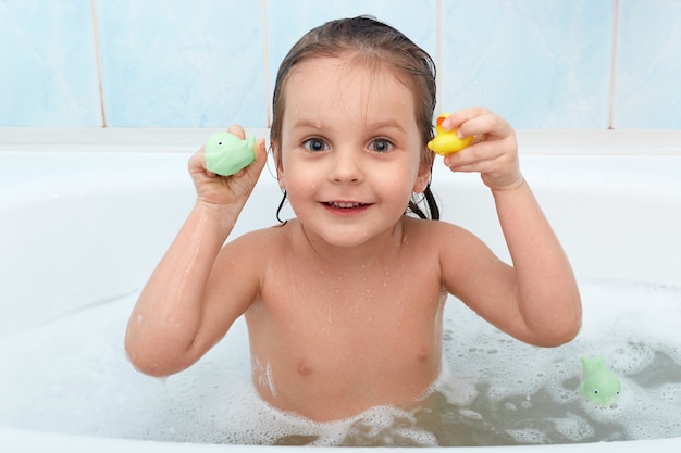 Garotinha segurando o brinquedo nas mãos tomando banho, brinca na água com pato e golfinho