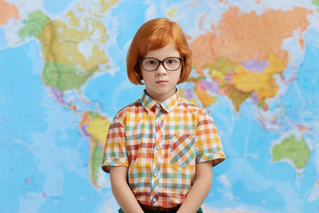 Foto grátis garotinho com cabelo ruivo, vestindo camisa quadriculada e óculos, de pé contra o mapa, indo para a escola. aluno inteligente em pé no gabinete de geografia na escola, vai ter lição