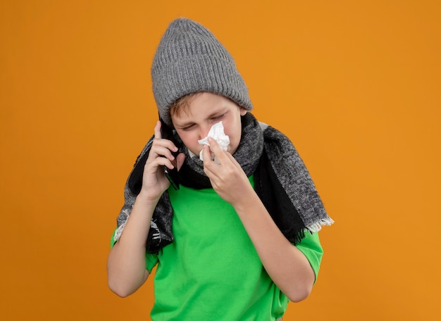 Garotinho doente, vestindo uma camiseta verde com um lenço quente e um chapéu, sentindo-se mal, falando no celular, limpando o nariz com um guardanapo de papel em pé sobre uma parede laranja