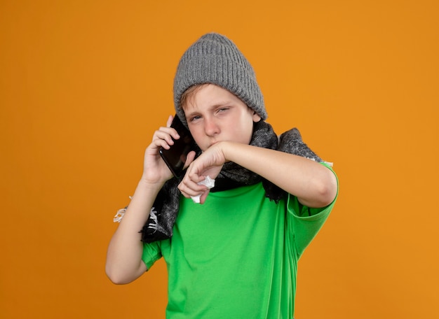 Garotinho doente, vestindo uma camiseta verde com um lenço quente e um chapéu, sentindo-se mal, falando no celular, limpando o nariz com um guardanapo de papel em pé sobre uma parede laranja