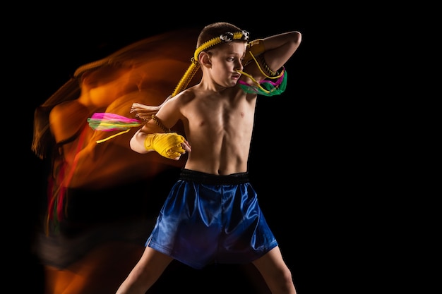 Foto grátis garotinho, exercitando o boxe tailandês na parede preta. luz mista. lutador praticando artes marciais em ação, movimento. evolução do movimento, momento de captura. juventude, esporte, conceito de cultura asiática.