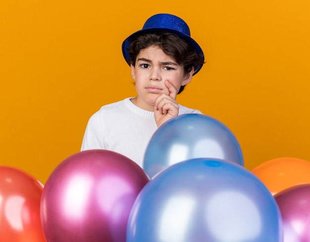 Foto grátis garotinho insatisfeito com chapéu de festa azul parado atrás de balões segurando o queixo