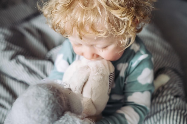 Garotinho louro e cacheado de pijama azul se senta na cama e brinca com seu brinquedo macio