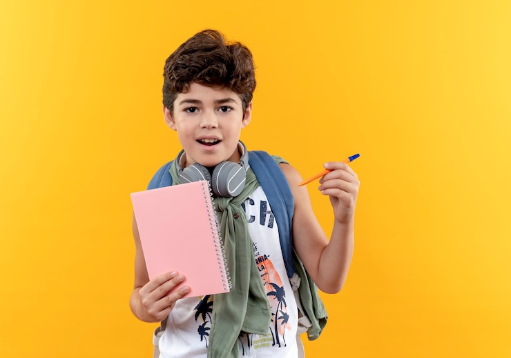 Foto grátis garotinho satisfeito com a mochila nas costas e fones de ouvido segurando um caderno com uma caneta