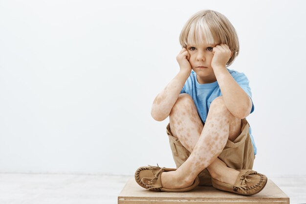 Garotinho se sentindo triste por não ser como todas as outras crianças. Criança loira fofa e infeliz sentada com os pés cruzados no chão, segurando o rosto com as mãos e olhando para o lado