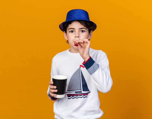 garotinho usando chapéu de festa azul soprando apito segurando xícara de café isolada na parede laranja