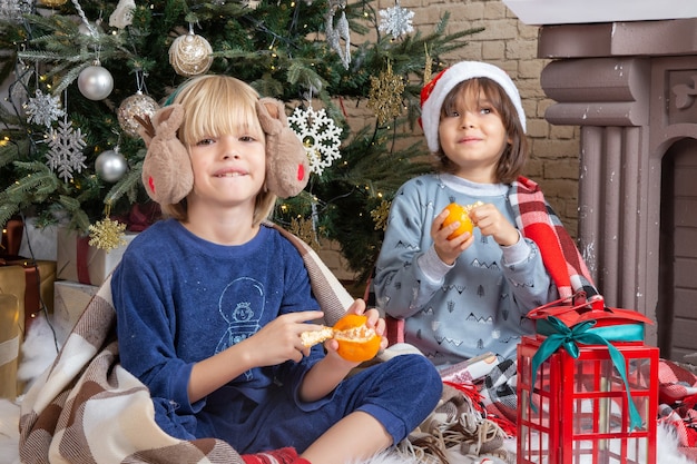 Garotinhos fofos sentados ao redor da árvore de natal e presentes em casa comendo garoto tangerina