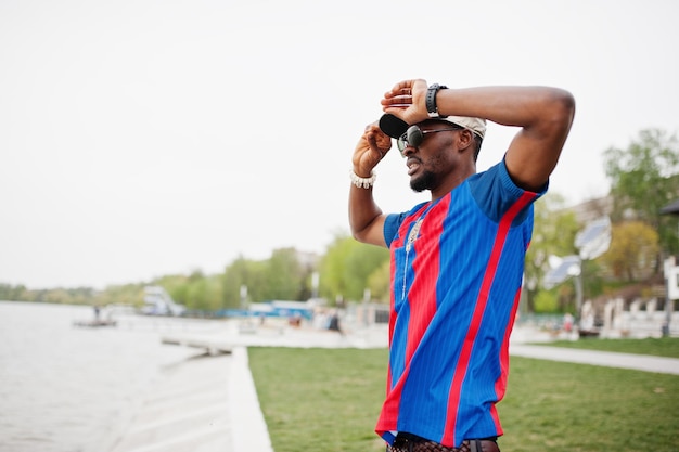 Garoto afro-americano elegante usa camiseta de futebol e óculos de sol ficam contra a praia do lago Retrato de homem esportivo negro