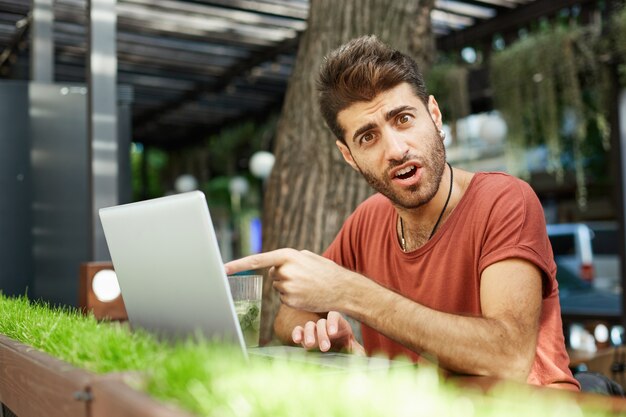 Garoto barbudo bonito confuso e surpreso fazendo perguntas sobre algo na tela do laptop, apontando para a tela com rosto maravilhado