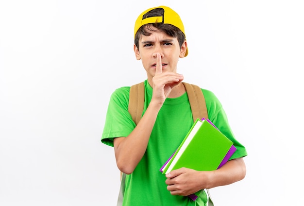 Garoto de escola rigoroso usando mochila com tampa segurando livros mostrando um gesto de silicone isolado na parede branca