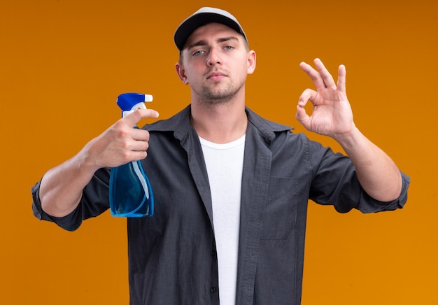 Garoto de limpeza bonito jovem confiante vestindo camiseta e boné, segurando o frasco de spray e mostrando um gesto de ok isolado na parede laranja