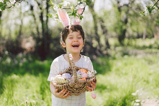 Garoto engraçado com cesta de ovos e orelhas de coelho na caça aos ovos de páscoa no jardim ensolarado de primavera