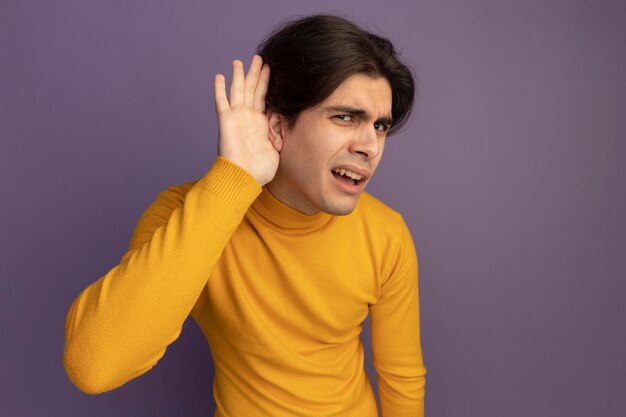 Foto grátis garoto jovem e bonito confuso, vestindo uma blusa de gola alta amarela, mostrando um gesto de escuta isolado na parede roxa