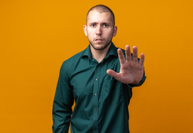 Garoto jovem e bonito descontente vestindo uma camisa verde e fazendo um gesto de pare com a mão